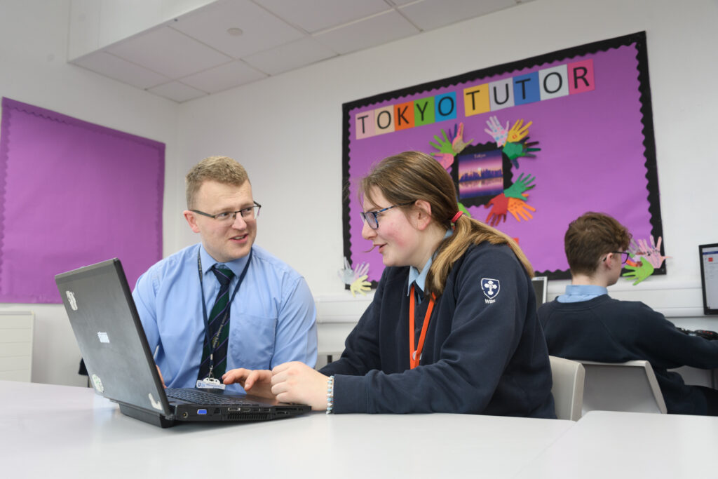 A female student working on a laptop smiling and a male teacher sat next to her smiling